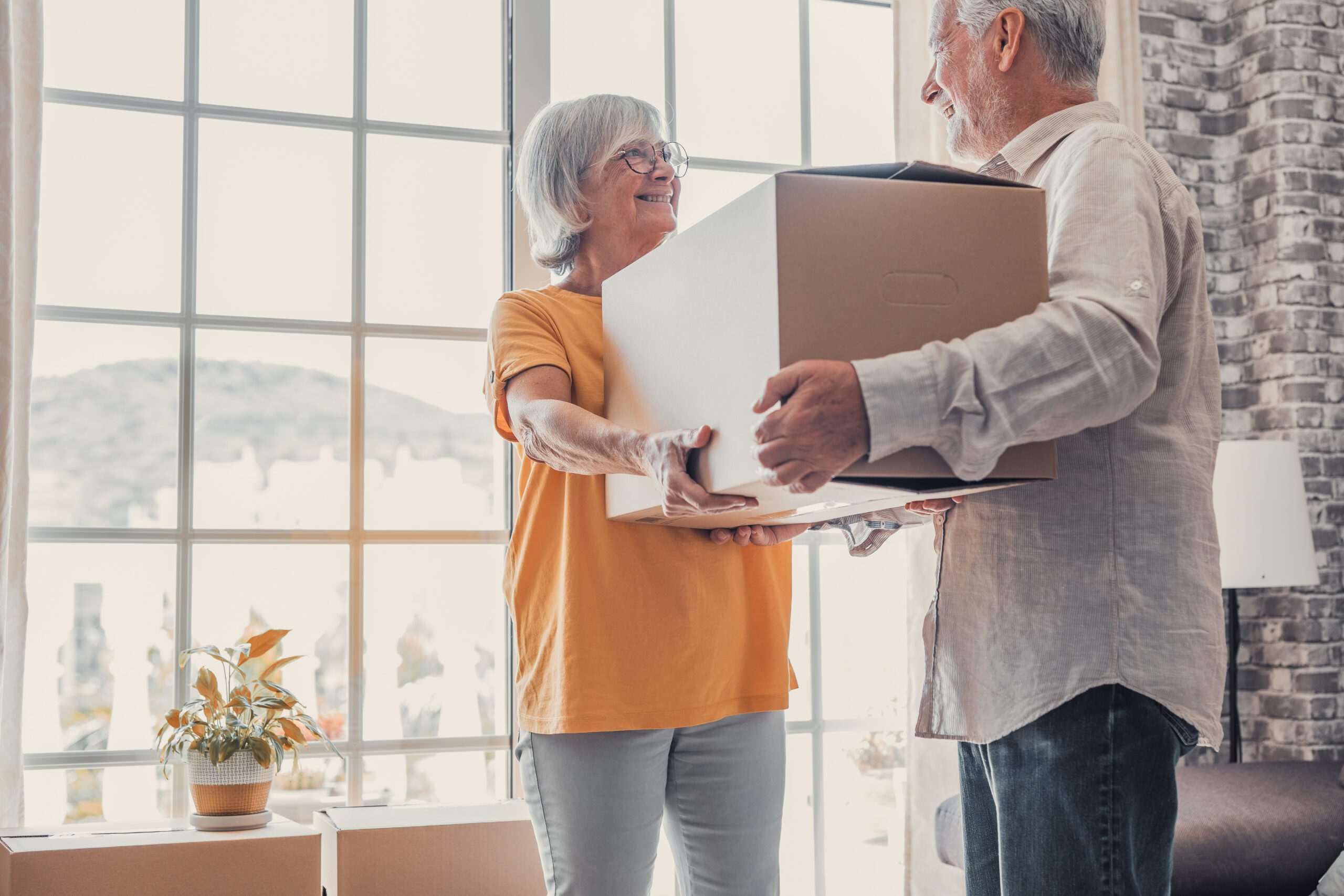 Older couple decluttering their space, carrying cardboard boxes into empty room with potted plants. Real estate property buying, relocation.
